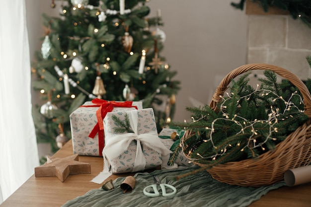 Presentes de Natal enrolados com estilo cesta rústica com galhos de abeto e decorações modernas contra árvore decorada festiva na sala escandinava Feliz Natal e Felizes feriados