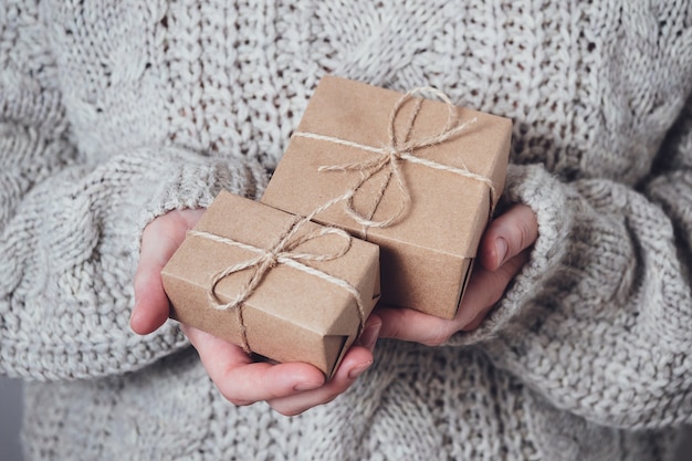 Presentes nas mãos das mulheres, close-up. Conceito de presente monocromático e minimalista. Uma garota com um suéter segura caixas de presente feitas de papel Kraft, amarradas com um barbante. Fundo surpresa, cartão de felicitações.