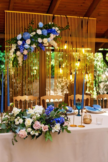 Presidium de bodas, mesa de banquete para recién casados con flores, vegetación, color azul y dorado. Arreglo floral exuberante en la mesa de la boda. Bombillas incandescentes. Enfoque selectivo suave.