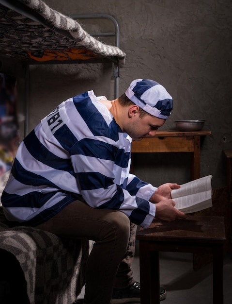 Preso con uniforme de prisión leyendo un libro o una Biblia mientras está sentado en una cama cerca de la mesita de noche con platos de aluminio en una celda de la prisión