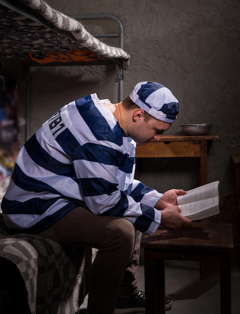 Foto preso con uniforme de prisión leyendo un libro o una biblia mientras está sentado en una cama cerca de la mesita de noche con platos de aluminio en una celda de la prisión