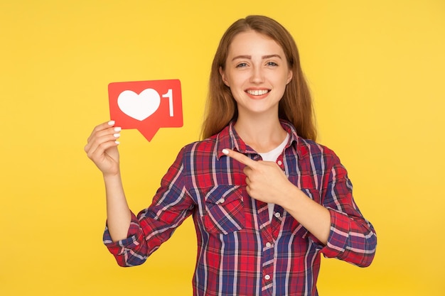 Pressione o botão do coração para desfrutar do conteúdo. Retrato de menina ruiva feliz em camisa quadriculada apontando para mídias sociais como ícone, símbolo de notificação de seguidor. tiro de estúdio interno isolado em fundo amarelo