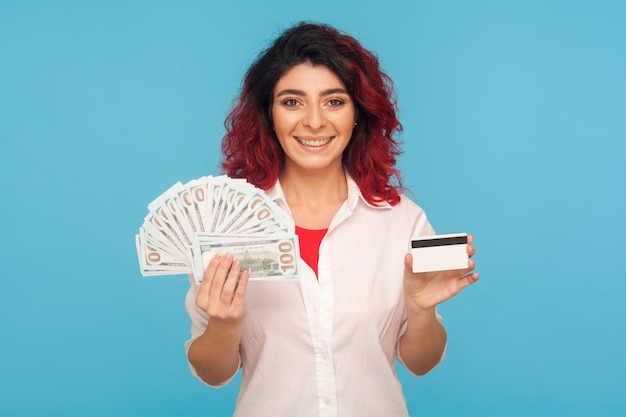 Préstamo bancario para compras, consumismo. Retrato de mujer hipster alegre con cabello rojo elegante con tarjeta de crédito, billetes de dólar y sonriendo complacido a la cámara. tiro del estudio aislado sobre fondo azul