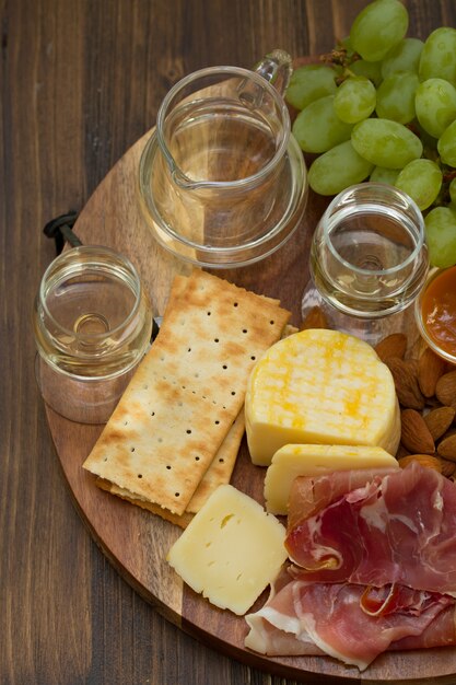 Presunto com queijo, biscoitos, uvas e vinho branco na mesa de madeira marrom