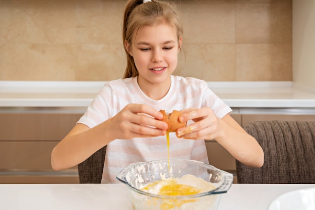 Preteen Mädchen bricht ein Ei in einer Schüssel auf einem Tisch und kocht in der Küche