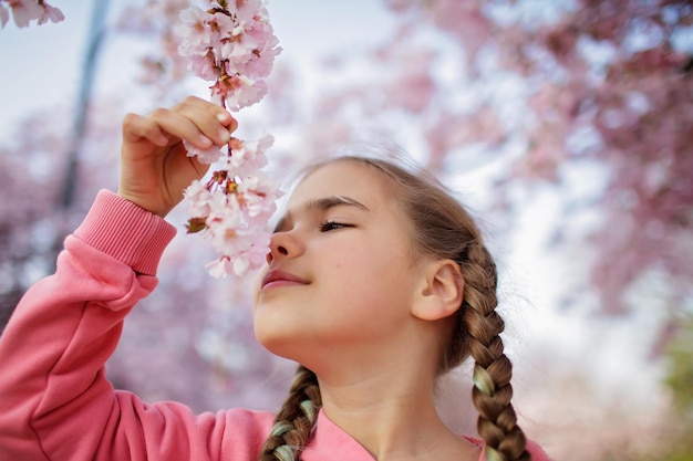 Preteen Mädchen riecht einen blühenden Sakura-Zweig, während sie im blühenden Garten spazieren geht, fröhliche Frühlingsstimmung