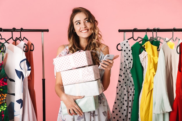 Pretty Woman con compra de pie en la tienda cerca de perchero y usando un teléfono móvil aislado en rosa