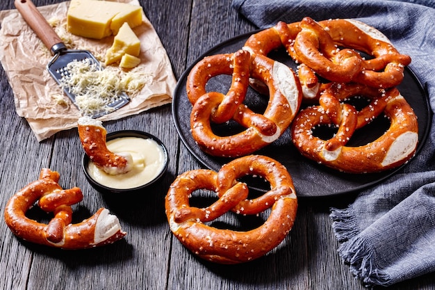 Foto pretzels macios recém-assados, assados em forma de nó e polvilhados com sal na chapa preta na mesa de madeira escura com molho de queijo, vista horizontal de cima