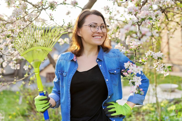 Primavera de jardinagem, retrato de mulher sorridente madura com ancinho. Árvores florescendo no fundo do jardim, limpeza sazonal no jardim, hobbies e lazer
