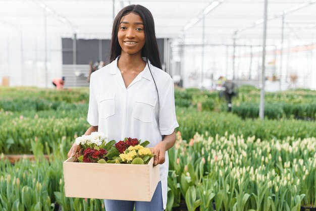 Primavera e mudas. Garota afro-americana sorridente com avental carrega uma caixa com plantas jovens em fundo de flores no interior da estufa