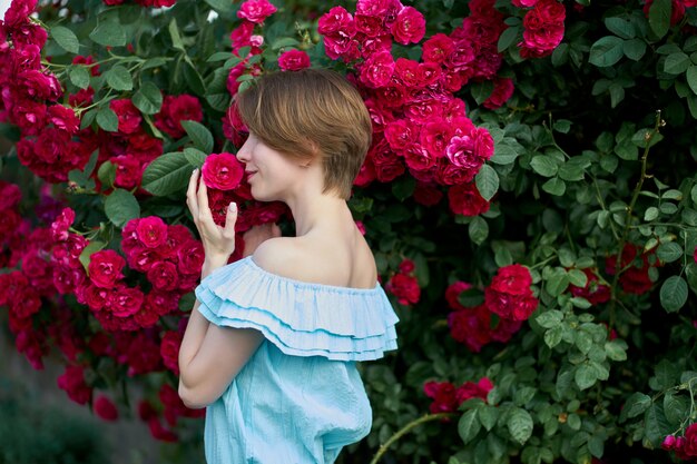 Primavera. Jovem mulher bonita do retrato que veste o vestido claro azul na moda que cheira rosas de florescência no jardim. Ao ar livre.