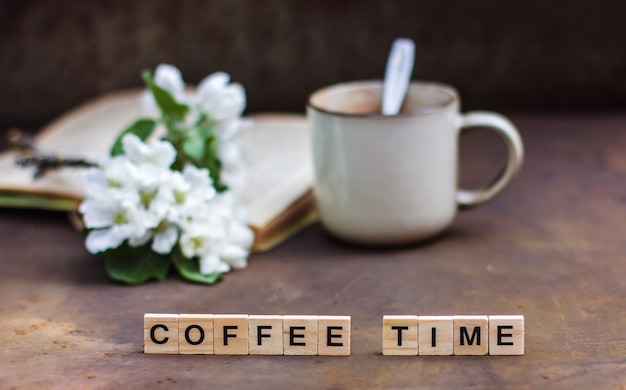 Primavera todavía la vida sobre un fondo oscuro. La inscripción "La hora del café" Una taza de café, un libro, una rama en flor de un manzano. Foto de estilo vintage, arreglo floral con rama