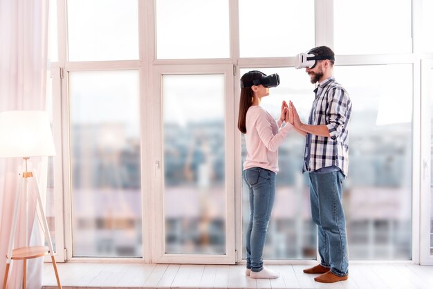 Primeira dança. Comprimento total de um casal doce e inocente que posa perto da janela enquanto experimenta os óculos de realidade virtual e se toca as mãos