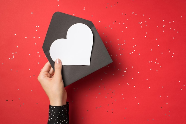 Primeira pessoa ver foto aérea de mão feminina segurando envelope preto com cartão branco de convite como coração dentro isolado no fundo vermelho com confete