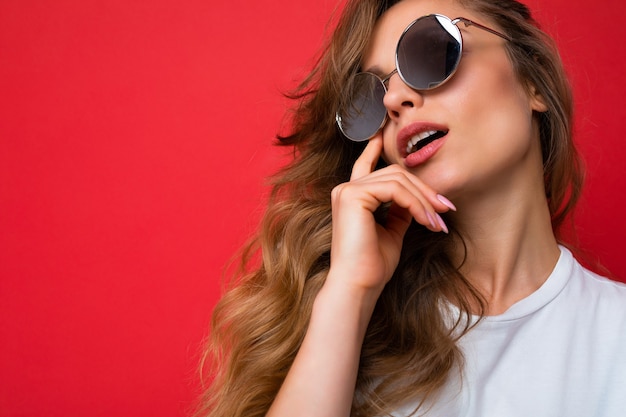 Primer disparo de la hermosa mujer rizada rubia oscura joven reflexiva sexy aislada sobre la pared de fondo rojo con camiseta blanca casual y gafas de sol con estilo mirando hacia arriba y pensando