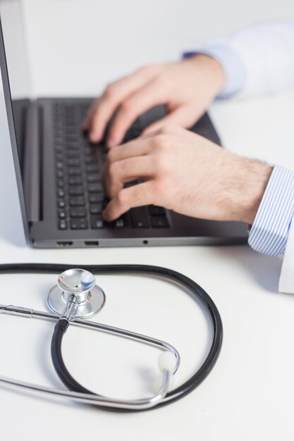 Foto primer del doctor que mecanografía en el ordenador portátil con el estetoscopio en el escritorio blanco