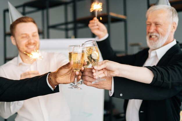 Primer equipo diverso de empleados multinacionales celebrando el éxito en la oficina sosteniendo luces de Bengala tintineando copas de champán y sonriendo alegremente en el Año Nuevo corporativo o Feliz Nochebuena