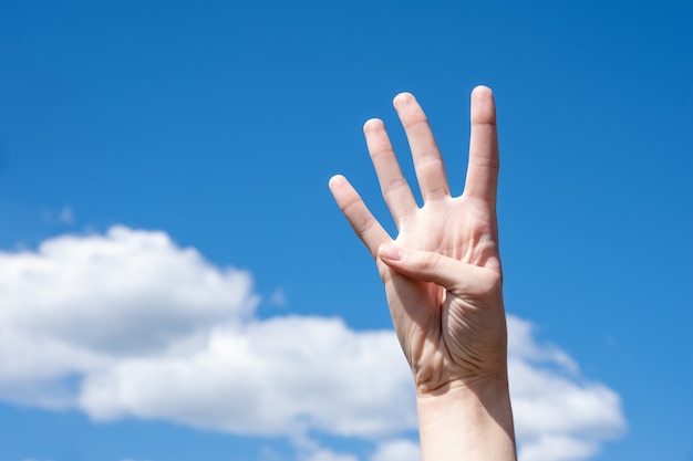 Primer gesto de la mano de una mujer mostrando cuatro dedos, aislado en un fondo de cielo azul con nubes, símbolo de lenguaje de señas número cuatro.