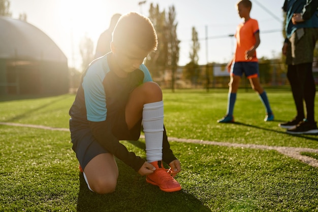 Primer niño pequeño jugador de fútbol atando cordones de botas de entrenamiento sentado en el campo de fútbol de hierba verde antes del partido