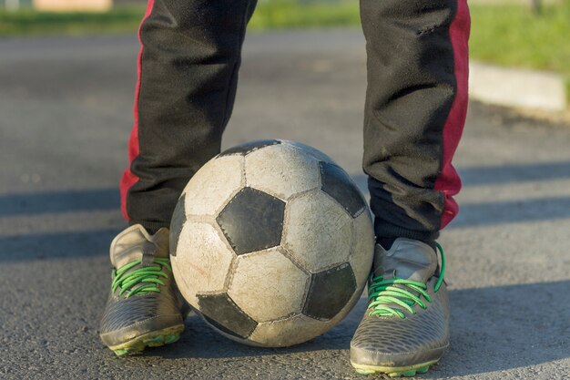 Primer de las piernas del niño en calzado deportivo que sostiene el balón de fútbol al aire libre en día soleado. Actividad de vacaciones para adolescentes, entrenamiento deportivo y concepto de recreación.