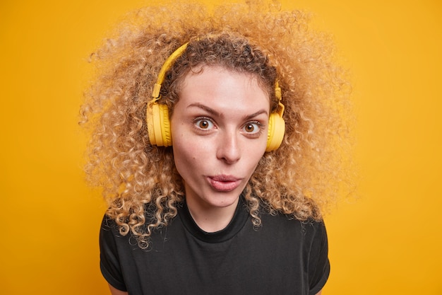 Primer plano de una adolescente rizada de buen aspecto usa auriculares inalámbricos en los oídos disfruta de la calidad del sonido escucha música mantiene los labios doblados vestidos con una camiseta negra informal aislada sobre una pared amarilla