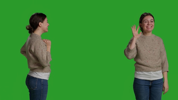 Primer plano de un adulto joven saludando a una persona con onda, sonriendo en el estudio. Mujer casual relajada saludando hola o hola en la cámara, de pie sobre el fondo de pantalla verde de cuerpo completo y diciendo adiós.