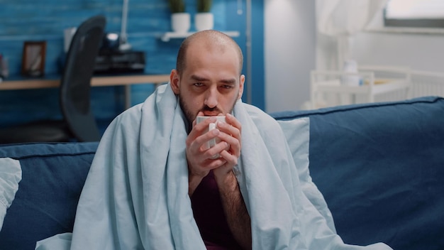 Primer plano de un adulto temblando y sosteniendo una taza de té contra los síntomas del virus. Hombre enfermo con escalofríos y temperatura en una manta mientras mira la cámara. Persona enferma con gripe y enfermedad.