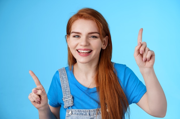 Primer plano alegre bailarina pelirroja despreocupada apuntando hacia arriba, levantando los dedos, sonriendo divertido, divirtiéndose, de pie fondo azul entretenido, disfrute de la fiesta musical, estado de ánimo disco, concepto de emociones despreocupadas