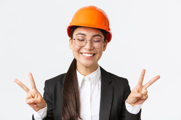 Primer plano de alegre ingeniera asiática exitosa mujer, arquitecto de la construcción en casco de seguridad y traje de negocios mostrando signos de paz y sonriente kawaii, fondo blanco de pie