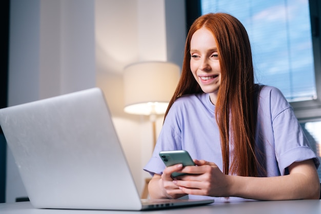 Primer plano, de, alegre, mujer joven, utilizar, teléfono, sentar escritorio, con, computadora de computadora portátil, en, sala