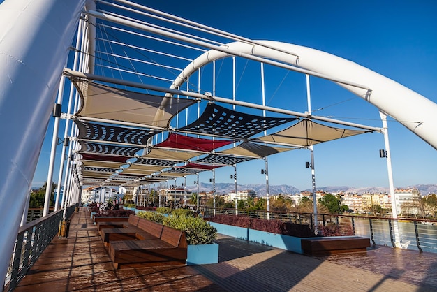 Primer plano de un amplio puente peatonal con áreas para sentarse, tumbonas y bancos bajo toldos del sol y hermosos macizos de flores verdes