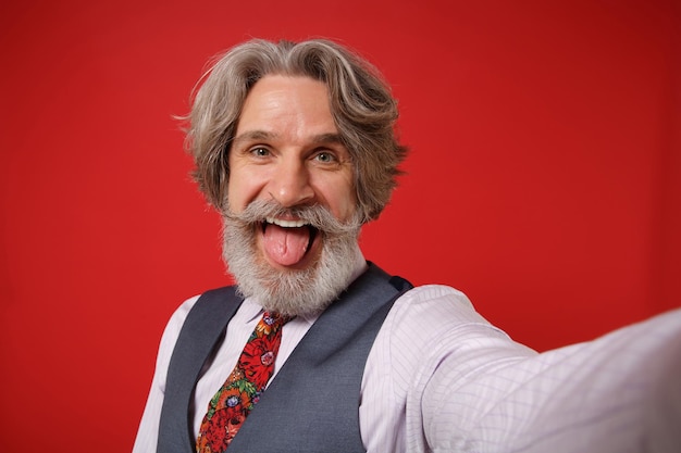 Primer plano de un anciano de pelo gris con barba y bigote, con chaleco de camisa y corbata, aislado de fondo rojo. Concepto de estilo de vida de las personas. Simulacros de espacio de copia. Haciendo selfie en el teléfono móvil, mostrando la lengua.