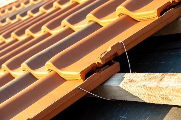 Primer plano de un anclaje de montaje metálico para la instalación de tejas cerámicas amarillas montadas sobre tablas de madera que cubren el techo de un edificio residencial en construcción.