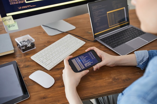 Foto primer plano de ángulo alto de manos femeninas sosteniendo el teléfono inteligente con código en la pantalla mientras trabaja en el escritorio en la oficina, concepto de desarrollador de ti femenino, espacio de copia