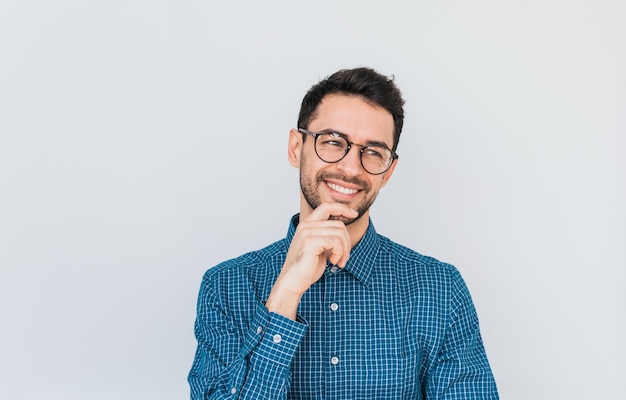 Primer plano de un apuesto empresario caucásico con gafas y camisa azul mirando hacia otro lado con una sonrisa feliz mostrando dientes blancos sosteniendo su barbilla Concepto de negocio y éxito de personas