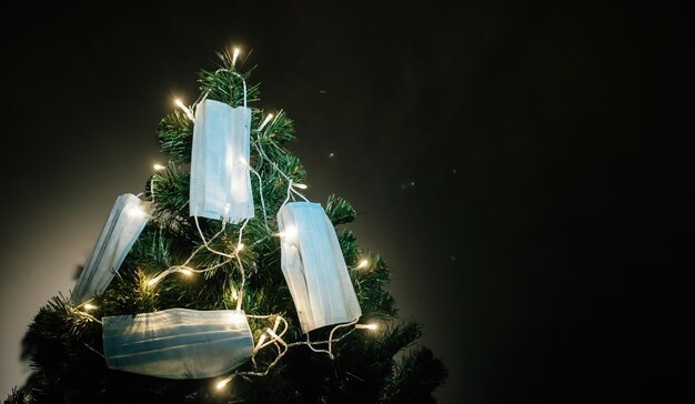 Primer plano del árbol de Navidad iluminado con guirnaldas, decorado con máscaras médicas protectoras en una habitación oscura. Ð¡a excepción de celebrar el año nuevo 2021 durante la pandemia de Covid-19. Copie el espacio.