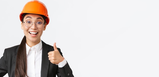 Primer plano de una arquitecta ingeniera jefa asiática complacida profesional en traje de negocios y casco de seguridad que muestra el pulgar hacia arriba en aprobación dar permiso recomendar empresa constructora