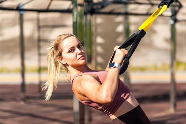 Primer plano atleta fuerte mujer rubia entrenando al aire libre tirando de las correas de suspensión trx haciendo ejercicios en la parte superior del cuerpo para desarrollar músculos quemar grasa mejorar el concepto de actividad deportiva y cuidado de la salud