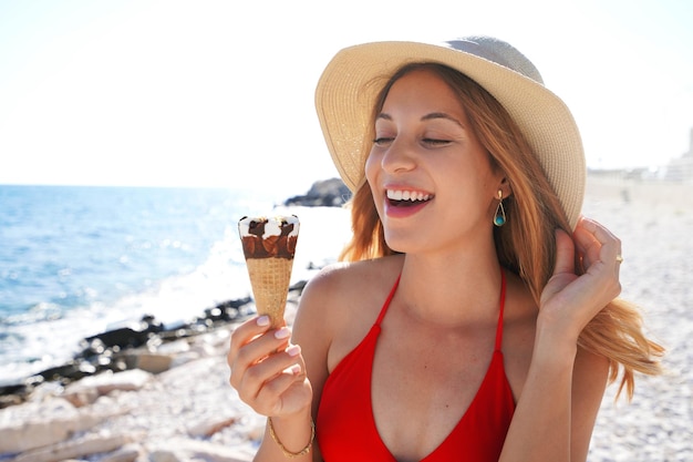 El primer plano de una atractiva mujer en bikini con sombrero sostiene y mira su cono de helado helado italiano en la playa en verano