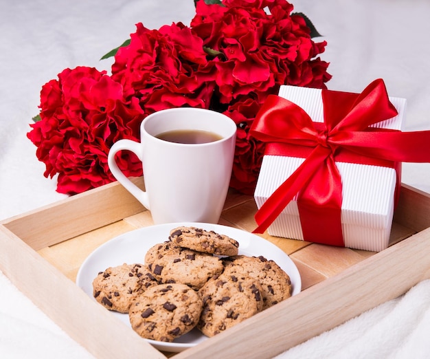 Primer plano de la bandeja de madera con galletas de chispas de chocolate, taza de té, flores y caja de regalo