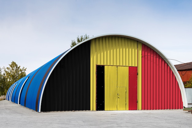 Primer plano de la bandera nacional de Bélgica pintado en la pared de metal de un gran almacén el territorio cerrado contra el cielo azul. El concepto de almacenamiento de mercancías, entrada a un área cerrada, logística.