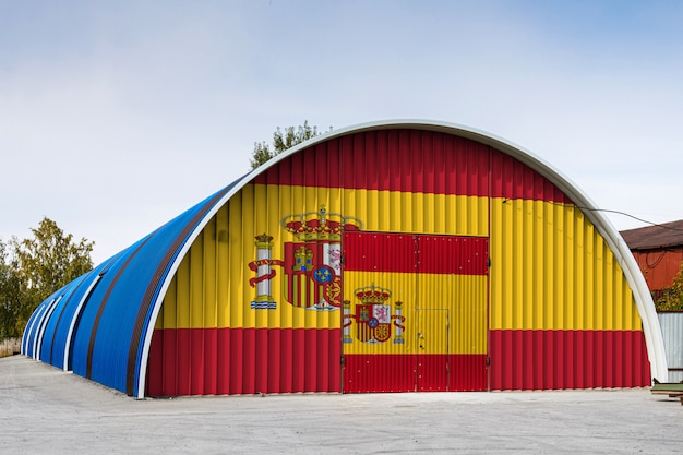 Primer plano de la bandera nacional de España pintada en la pared de metal de un gran almacén del territorio cerrado contra el cielo azul. El concepto de almacenamiento de mercancías, entrada a un área cerrada, logística.