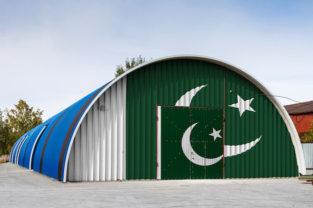 Primer plano de la bandera nacional de Pakistán pintado en la pared de metal de un gran almacén del territorio cerrado contra el cielo azul.