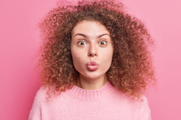 Foto primer plano de una bella joven europea rizada mantiene los labios doblados y ha sorprendido la expresión de la cara expresa que el amor usa un suéter quiere besarte aislado sobre una pared rosa. mwah para ti