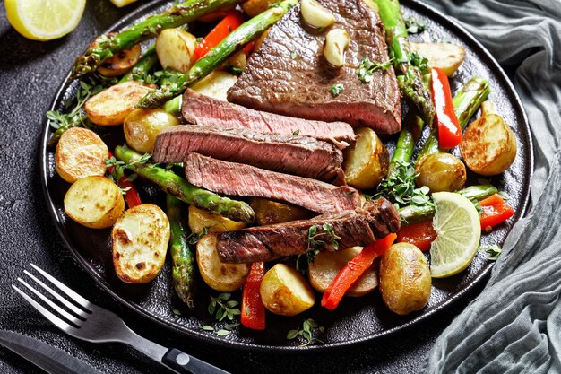 primer plano de bistec de ternera con verduras asadas patatas nuevas, espárragos y pimiento rojo en un plato negro