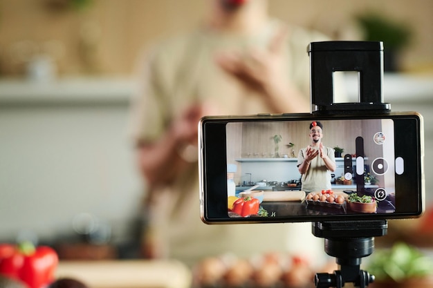 Foto primer plano de un blogger que filma con un teléfono inteligente lanzando un tomate mientras está de pie cerca de la mesa de la cocina