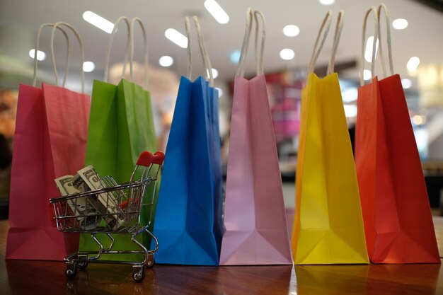 Primer plano de bolsas de compras coloridas con monedas de papel en un carrito de compras en miniatura en una mesa de madera