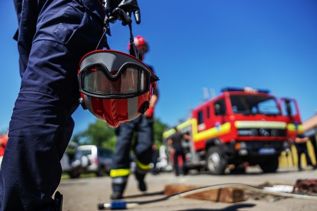 Primer plano de bombero sosteniendo su casco y caminando hacia el camión de bomberos.