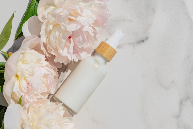Primer plano de una botella blanca con crema cosmética natural y flores de peonía rosa florecientes sobre un fondo de mármol blanco Concepto de belleza cosmética Espacio de copia