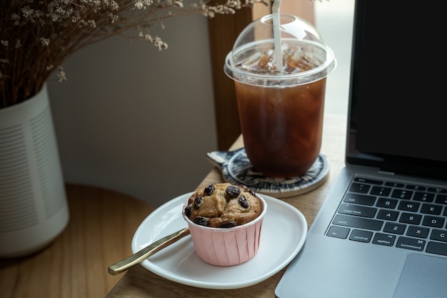 Primer plano de café helado en taza taza y pastel de taza de plátano casero con teclado computadora portátil en escritorio de madera escritorio de oficina en cafetería en la cafetería, durante el concepto de trabajo empresarial